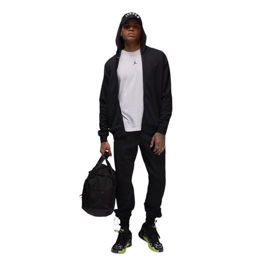 Person wearing a black tracksuit with a white shirt, holding a black bag on a white background